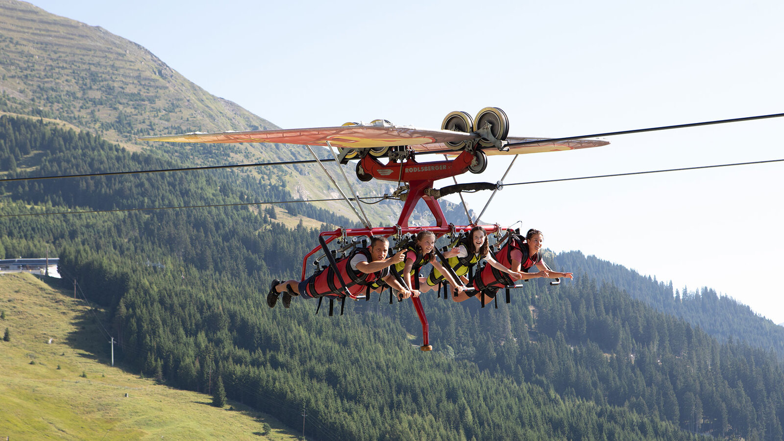 Mehrere Personen fliegen angeschnallt an einer Seilkonstruktion über eine Berglandschaft.
