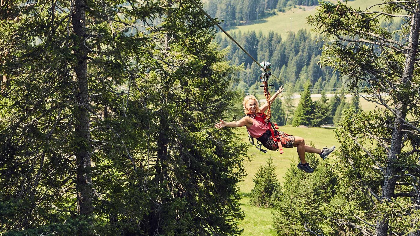 Person fährt mit einer Seilrutsche durch einen alpinen Wald mit Blick auf grüne Wiesen.