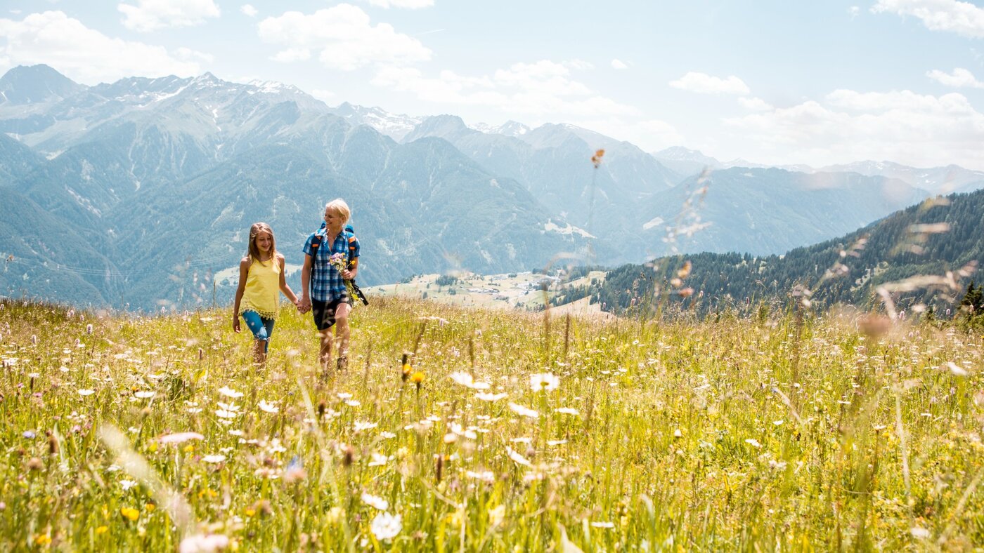 Mutter und Tochter gehen Hand in Hand über eine Blumenwiese mit Bergpanorama.