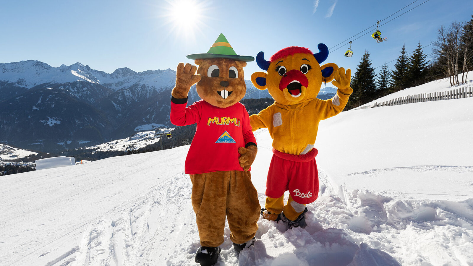 Zwei verkleidete Maskottchen stehen auf einer verschneiten Skipiste in den Bergen und halten die Arme in die Höhe.